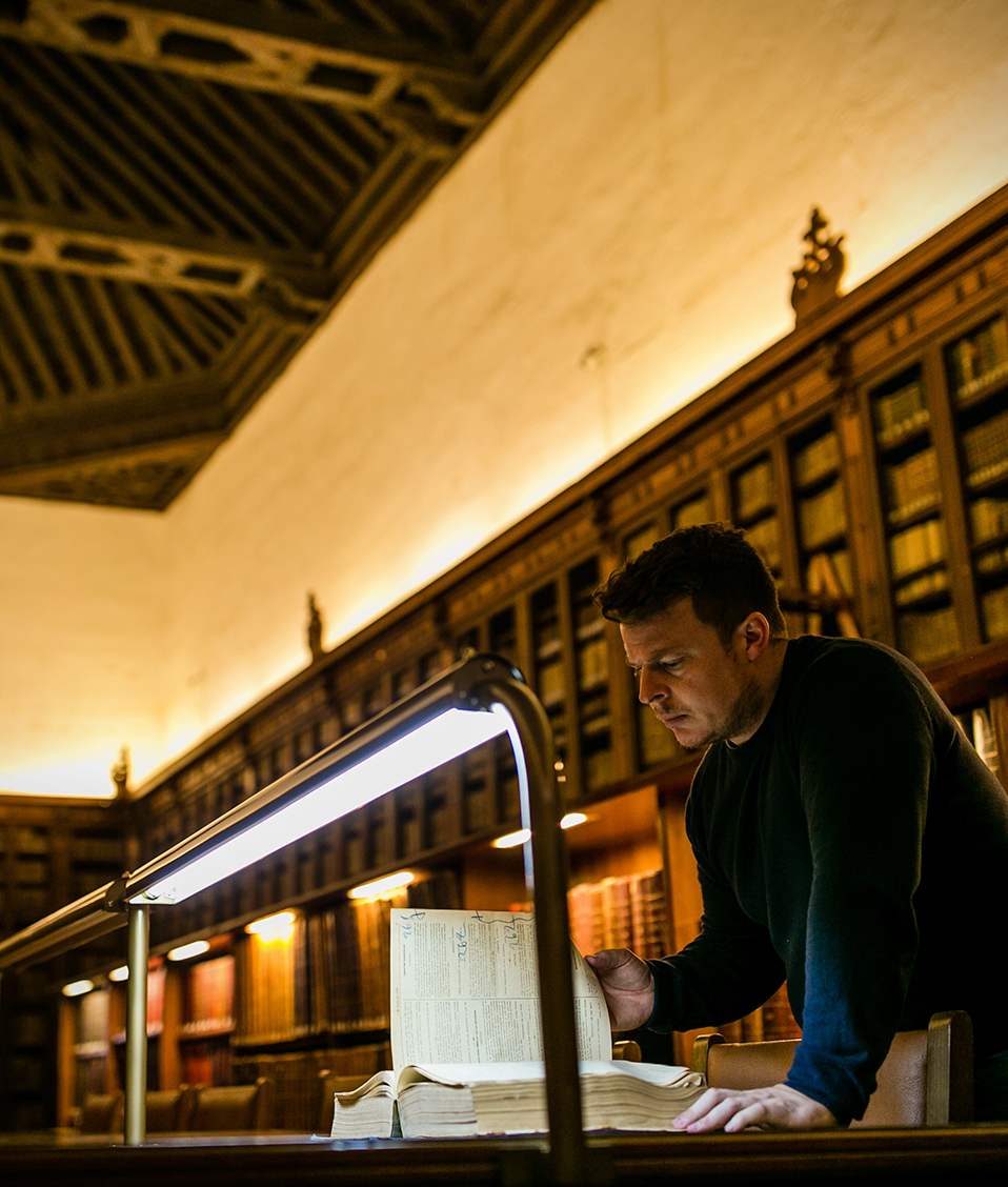Persona leyendo un libro antiguo en la biblioteca del Hospital Real con puerta al fondo con escudo antiguo UGR